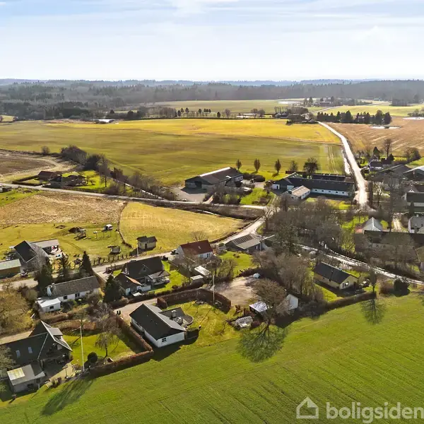 En landsby set ovenfra med spredte huse. Veje forbinder husene, der omgives af åbne marker og skove i baggrunden. Der er en klar, blå himmel uden skyer.