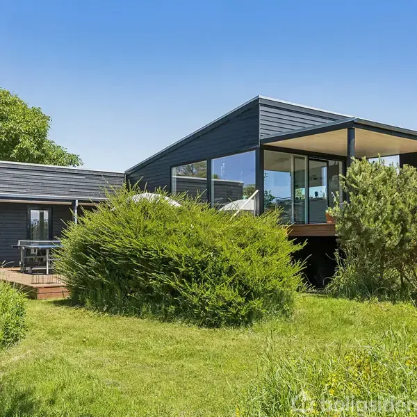 Sort moderne hus står på græsplæne, omgivet af buske og træer under en blå himmel. Huset har store glaspartier og en udendørs terrasse.