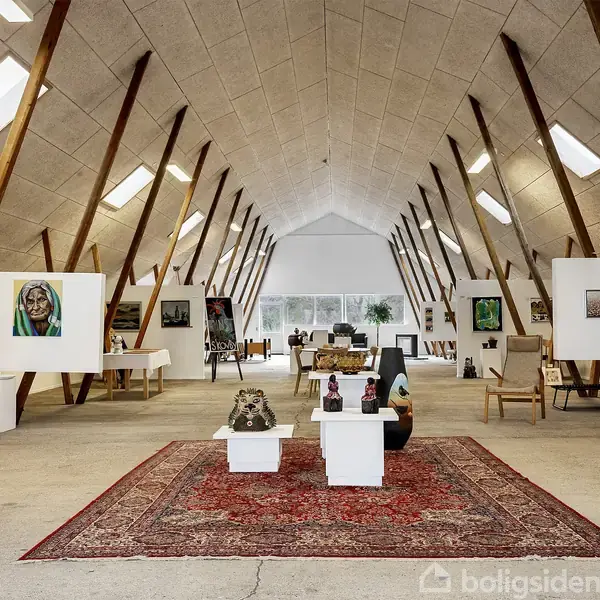 Et kunstgalleri med malerier og skulpturer udstillet. Malerier hænger på væggene og nogen står på gulvet. Rummet er stort og lyst med et tæppe midt på gulvet.