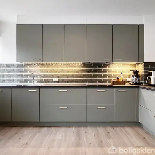 Gråt køkken med overskabe, skuffer, og sorte bordplader. Underbelysning lyser backsplash af mørke fliser. Til venstre et vindue med planter; til højre en kaffemaskine. Lys trægulv.