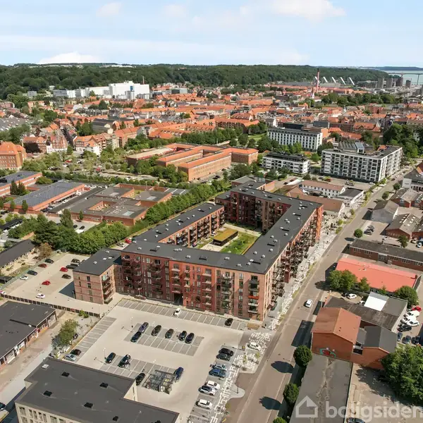 Rød murstensbygning med indre gårdhave står stille blandt andre bygninger. Parkeringsplads foran bygningen. Byen og skovklædt bakke udgør baggrunden.