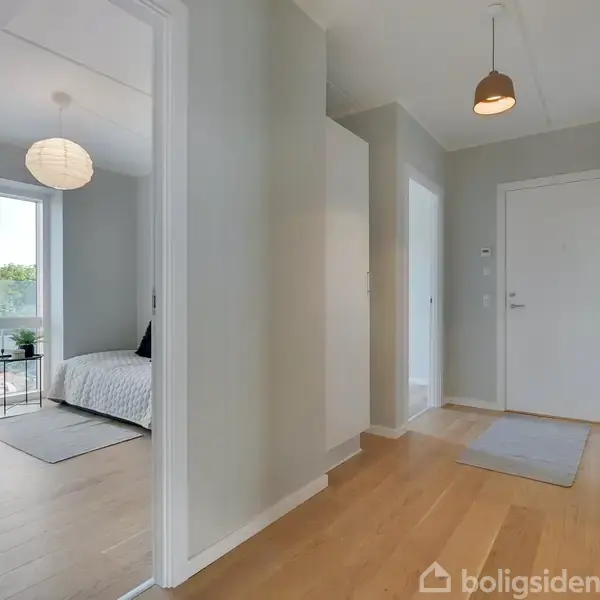 Gangområde med lyst trægulv og hvide vægge. Til venstre ses et soveværelse med en seng og stort vindue. Dæmpet indretning med moderne lamper og minimalistisk design.
