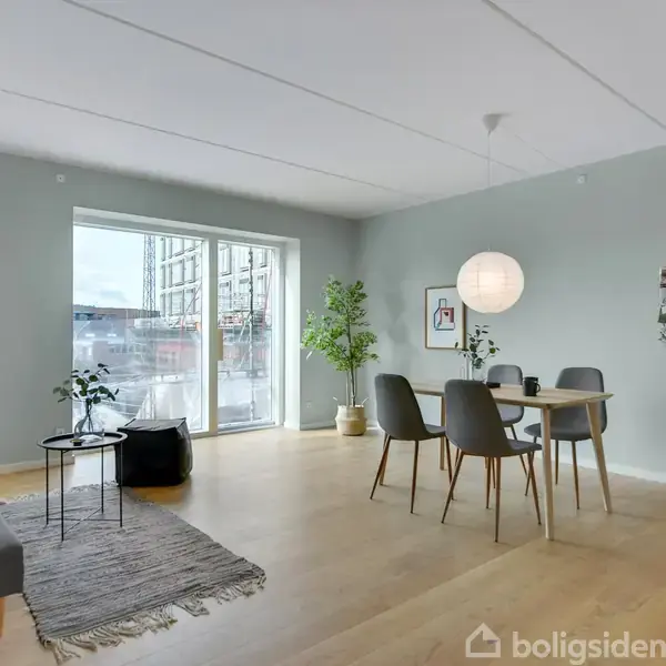 Gråt sofaområde med bord og tæppe, står i en lys stue. Spisebord med fire stole og planter omkring, ved et stort vindue. Lyse trægulve og vægdekorationer skaber en moderne atmosfære.