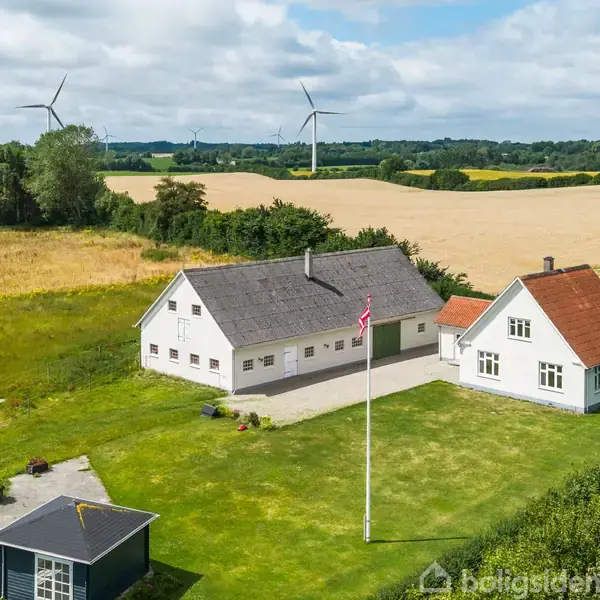Et lyst hus med gråt tag står på en velplejet græsplæne med flagstang, omgivet af marker og træer. I baggrunden ses vindmøller under en delvist skyet himmel.