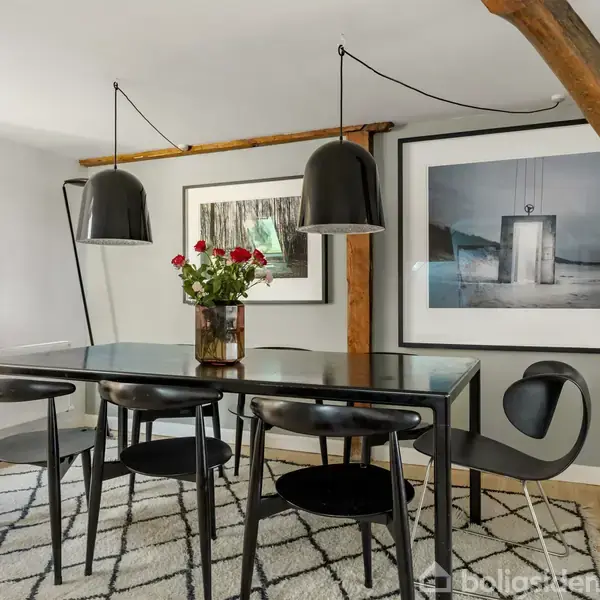 Et spisebord i sort med seks stole står på et mønstret tæppe. To sorte lamper hænger over bordet. En vase med røde blomster er placeret centralt. Baggrunden har kunst på væggen.