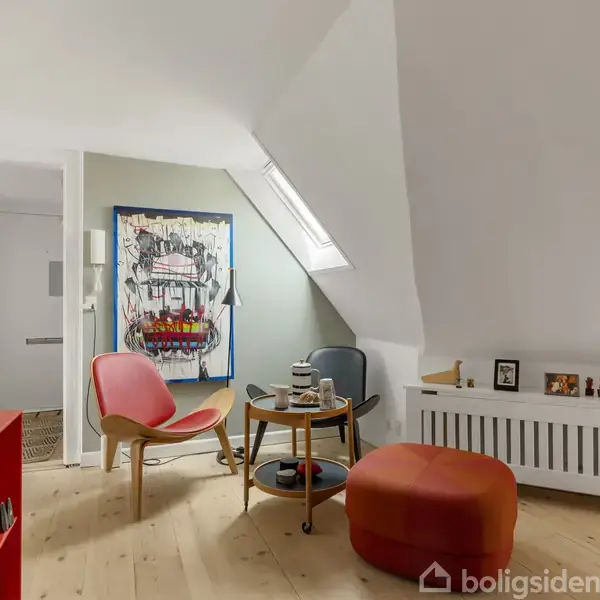 Et moderne stueområde med en rød lænestol og orange puf omkring et lille rundt bord. Lyse trægulve, kunst på væggen og skråloftsvindue skaber en lys atmosfære.
