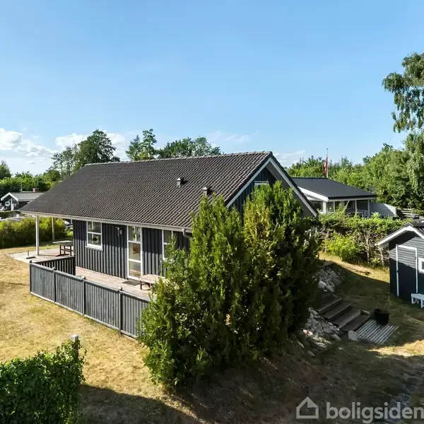 Et gråt hus med terrasse står omgivet af grønne træer og buske i en velplejet have. Ved siden af huset er et lille skur. Himlen er klar og blå.
