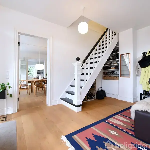 En entré med en trappe der fører opad, omgivet af hængende overtøj og sko på en rack. En dør fører til en spisestue med træmøbler. En plante står ved siden af døren.