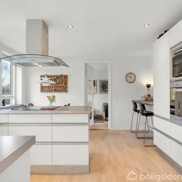 Køkken med hvidt moderne design, åbent til spisestue. En køkkenø står centralt med emhætte. Der er også indbyggede apparater, barområde med høje stole og vægur. Naturligt lys strømmer ind gennem vinduerne.