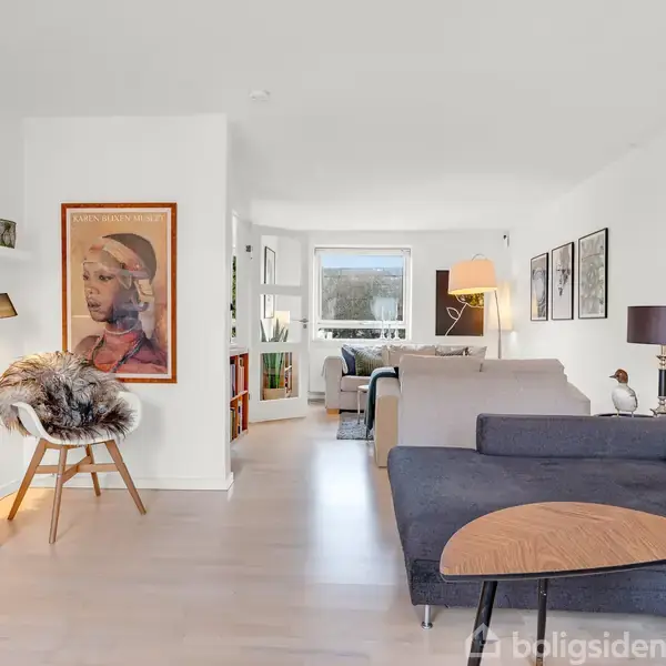 Stue med moderne møblering, hvor en grå sofa står centralt. På væggen hænger en plakat og hylder med pynt. I baggrunden ses et vindue og en bogreol.