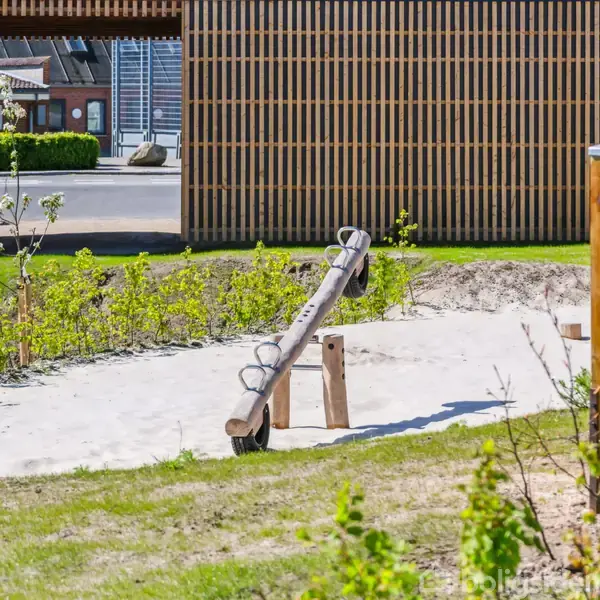 En vippe står ubenyttet i en sandkasse i en park. Omkring vippen er der grønne buske og en træstruktur, som danner en baggrund af trælameller.