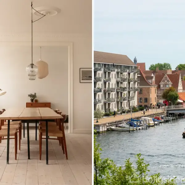 Spisebord med otte stole står i lyst rum med hængende lamper. Ved siden af ses en byudsigt over en flod med både, omgivet af farverige bygninger.