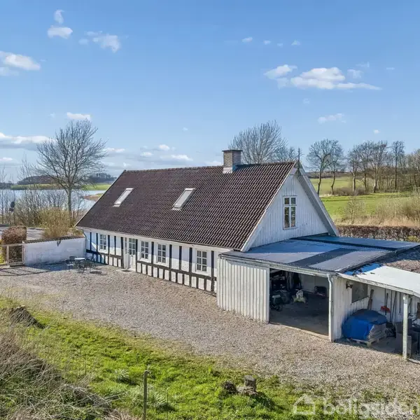 Et hvidt bindingsværkshus med mørkt tegltag står på en grusbelagt grund. Ved siden af er en carport. I baggrunden ses en sø og træer under en blå himmel.