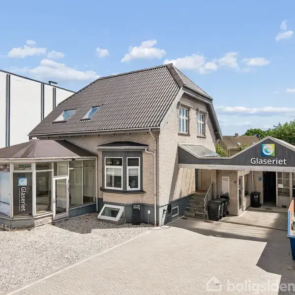 Et hus med skiltet "Glaseriet" hængende ved indgangen i en gård, på en solrig dag. Bygningen har flere vinduer og et gråt tag, omgivet af en flisebelagt indkørsel.