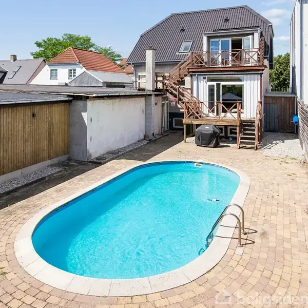 En oval swimmingpool ligger i en gårdhave omgivet af en stenbelagt terrasse. I baggrunden er et halvandenplanshus med trætrappe op til en terrasse med udsigt over poolen.