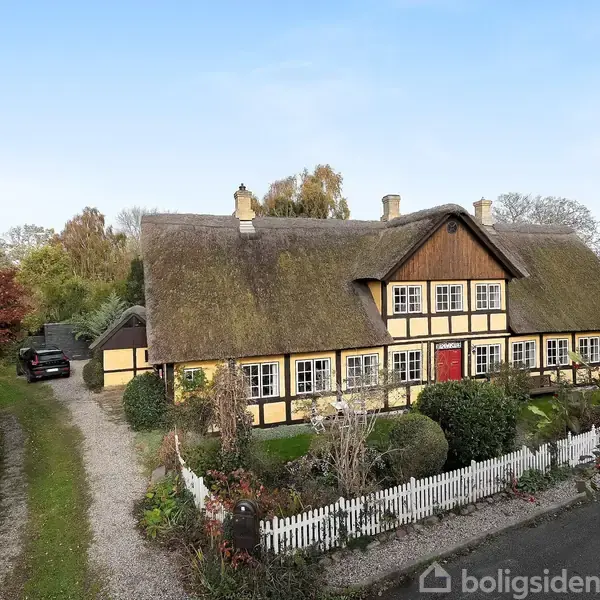 Bindingsværkshus med stråtag står i en grøn have omgivet af buske og træer. Husets gule facade med hvide vinduer er fremtrædende, og taget har flere skorstene.