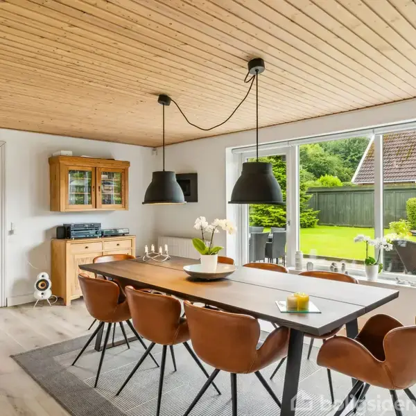 En lang spisebord med brune stole står i en lys spisestue. To lamper hænger over bordet. Vinduer viser en grøn have udenfor. En skænk står i baggrunden.