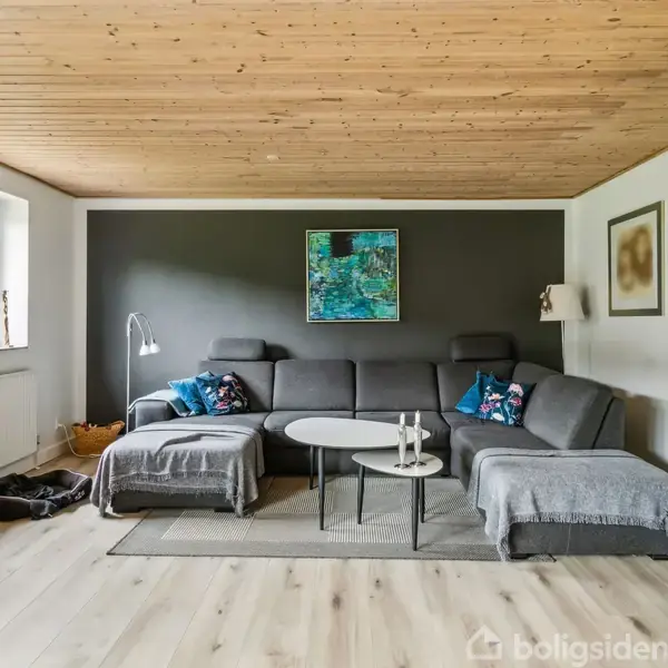 En grå hjørnesofa står i en stue med træloft, placeret mod en mørk væg. To små borde og farverige puder bidrager til indretningen, mens lys strømmer ind fra vinduet.