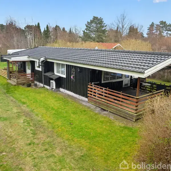 Sort træhus med overdækket terrasse står stille på en grøn græsplæne omgivet af buske og træer. Et skilt med teksten "Ny bolig" er placeret i forgrunden.