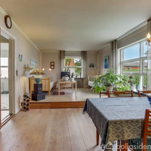 En spisestue med et træbord dækket af en mønstret dug; omgivet af træstole, husplanter ved vinduet; bogreoler og vægkunst skaber en hyggelig atmosfære i et lyst rum.