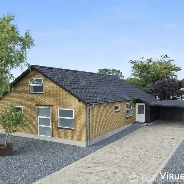 Et gult murstenshus med sort tag står stille ved siden af en bred indkørsel, der fører til en carport. En grusbelagt have omgiver huset. Tekst: "Visuelt forslag".