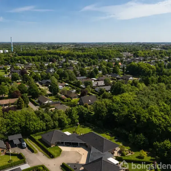Billedet viser et boligområde set fra luften, hvor huse med haver omgivet af grønne træer er spredt ud i landskabet. Det er solskin, og himlen er klar.