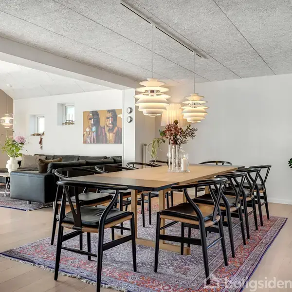 Spisebord med sorte stole står på et tæppe, omgivet af en stue med sofa, pejs og kunstværk på væggen. Lysekroner hænger over bordet i et moderne rum.
