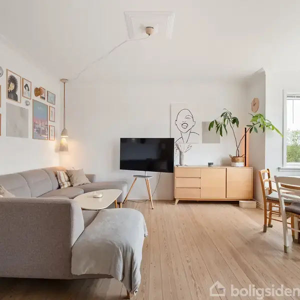 En lys stue med en grå sofa og et sofabord til venstre. Et TV står på en stander foran en kommode. Kunstværker pryder væggene. En spiseplads ved et vindue til højre. Plante ved siden af kommoden.