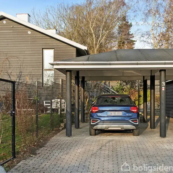 Bil parkeret under en carport ved siden af et moderne, træbeklædt hus. Omkring er der en brolagt indkørsel, et hegn og vintertræer. Synligt husnummer: 31.