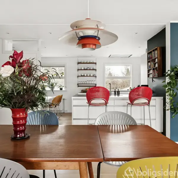 En spisebord med farverige stole og en blomsterdekoration i forgrunden. I baggrunden ses et moderne køkken med røde barstole, planter og vægdekorationer.