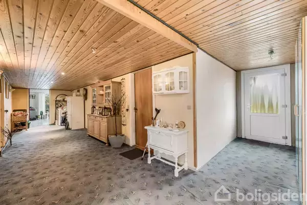 Gang med træloft, tæppebelagt gulv og møblering med skabe og kommoder. I venstre side er en dør, mens glasdøren med matteret rude er i højre endevæg.