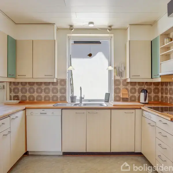Et L-formet køkken med hvide skabe og træbordplader. En vask står under et vindue, omgivet af lys beige fliser. En ovn og en kedel er i højre side.