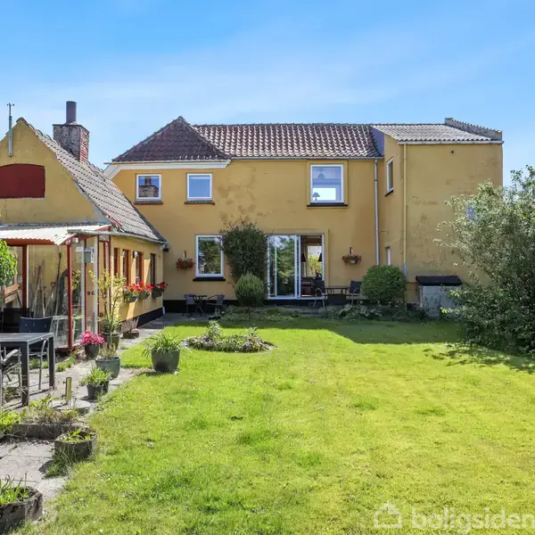 Gul villa med to etager står i en velplejet have. Huset har et skifertag, mange vinduer og en overdækket terrasse med havemøbler. En græsplæne strækker sig hen til huset.