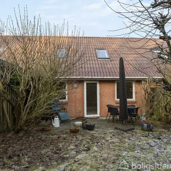 Et rækkehus ses med en terrasse, hvor der står et bord og stole. I haven omkring er der nøgne træer og buske. En mørk parasol er centralt placeret.