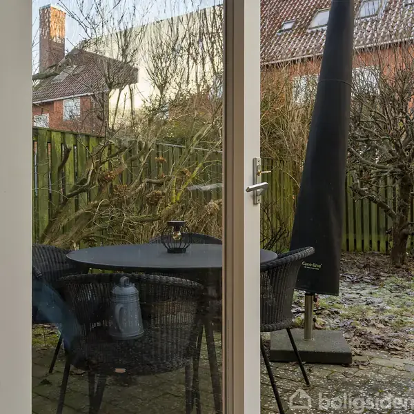 En trægårdsdør viser en terrasse med et rundt bord og kurvestole omkring det. En lanterne står på bordet, og en sort parasol står ved siden af. Baggrunden er en have med buske og et hegn.