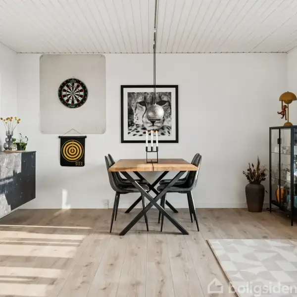 Et spisebord med fire stole står på et trægulv i et lyst rum. På væggen hænger to dartskiver og flere billeder. En glasvitrine og dekorative genstande pryder rummet.