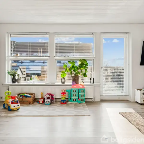 Legetøj står stille i en lys stue med store vinduer. Rummet har en plante, en legetøjsbil, og et dukkehus, der skaber en indbydende og barnlig atmosfære.