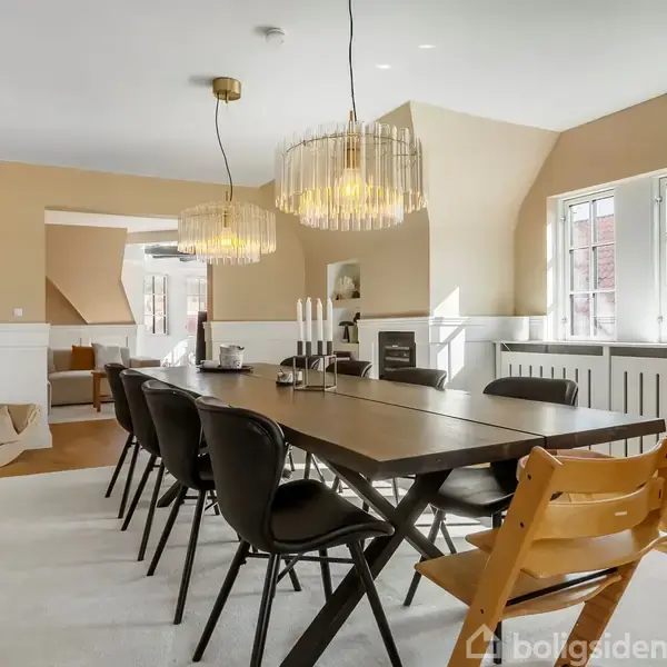 Et langt spisebord med sorte stole står i en elegant spisestue. Over bordet hænger to moderne lysekroner. Rummet har lyse vægge, store vinduer og smukt trægulv.