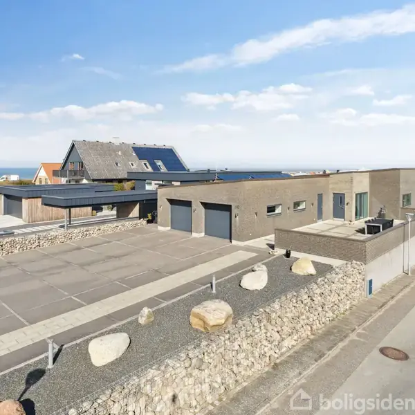 Et moderne, minimalistisk hus med fladt tag og store vinduer står på en velplejet grund af sten og fliser. Huset ligger i et kystnært område med flere omkringliggende huse.