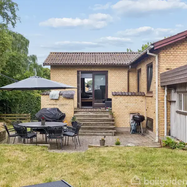 En terrasse med et bord og stole under en parasol ved siden af en gul murstensbygning. Der er en grill nær trappen, omkranset af græs og buske under en blå himmel.