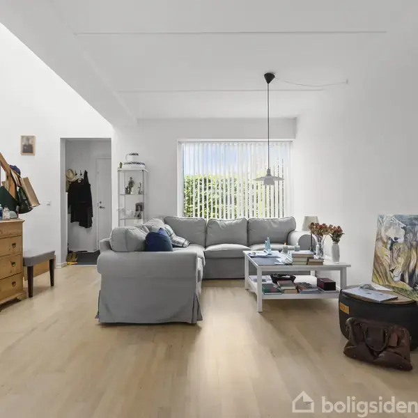 Grå sofa står midt i en lyst, moderne stue med trægulve. Et sofabord er placeret foran sofaen, omgivet af en kommode og malerier. Vindue bagerst giver naturligt lys.