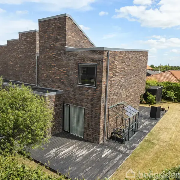Et modernistisk, murstenshus med fladt tag står i en velplejet have. Det har en glasdør mod en terrasse og en lille glaspavillon ved siden. Omgivet af grønne hække og plæner.