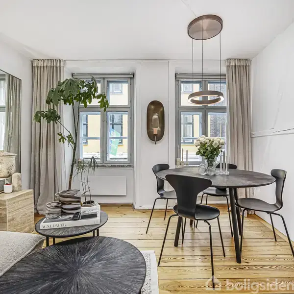 Et moderne spisebord med fire stole står i en lys stue. Et stort vindue med gardiner bagved giver naturligt lys. Indretningen inkluderer en plante og dekorative genstande.