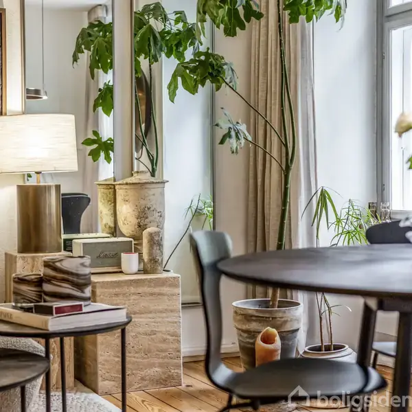 En elegant stue med en rund spisebord, omgivet af potteplanter og store vinduer. En lampe lyser op ved siden af en sofa, mens bøger ligger på et sidebord.