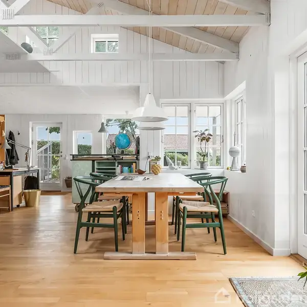Et lyst spisestueområde med et træbord omgivet af seks grønne stole. Rummet har store vinduer, der slipper masser af lys ind, og synlige loftsbjælker. En plante og en lampe pynter bordet.