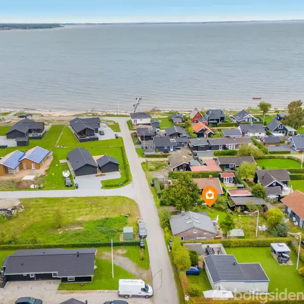 Boligområde tæt ved kysten omgivet af græsplæner og huse. Havet strækker sig i baggrunden, mens veje og biler ses blandt bygningerne. Husene har forskellige farver og indretninger.