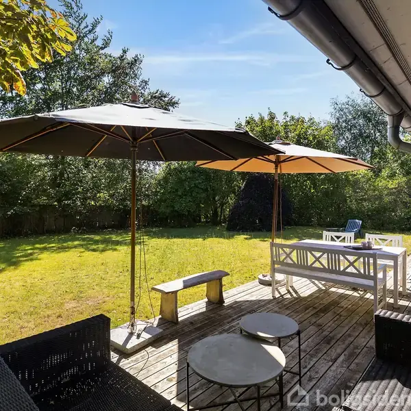 To sorte parasoller står åbne på en træterrasse med havemøbler. Baggrunden er en frodig have med græsplæne og træer under en klar blå himmel.