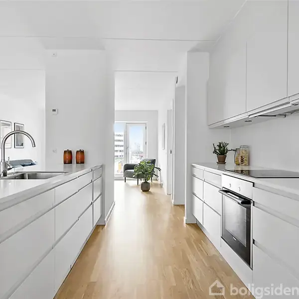 Køkken med hvide skabe og moderne apparater. En vask med høj vandhane på en ø. Trægulv fører til lys stue med planter.