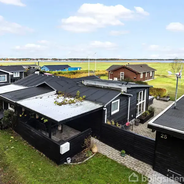 Sort sommerhus med fladt tag og overdækket terrasse; står i en række af lignende huse med udsigt over en grøn mark og kystlinje i baggrunden. Nummeret "25" ses på huset.