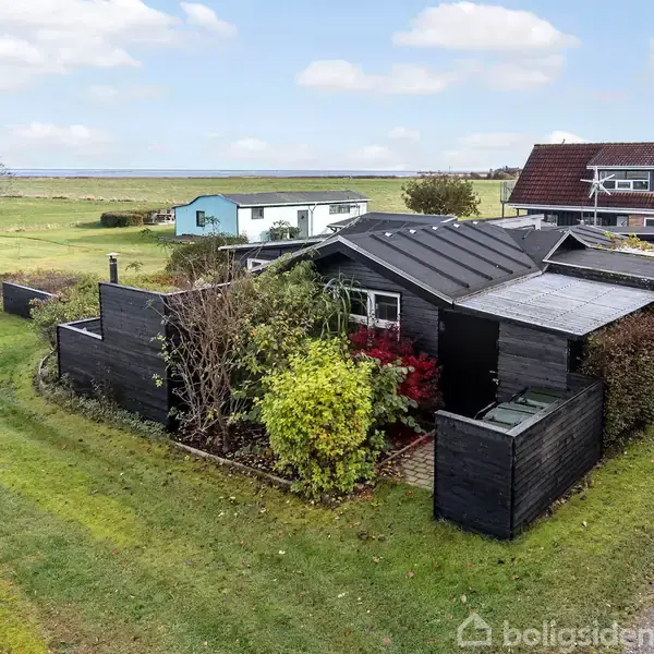 Sort hus omgivet af tæt løv og græsplæne, placeret i et åbent landområde med flere huse i baggrunden og en blå himmel over. En rød bil holder parkeret til venstre.